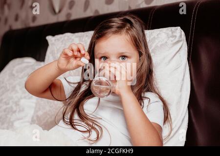 a sick child, a girl sitting on a bed with a white pillow, drinking water from a glass and holding a white pill in hand Stock Photo