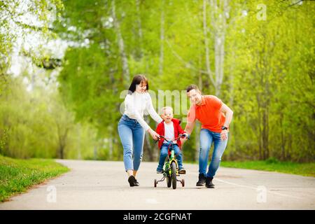 Dad and mom teaches little son to ride bike in park, have fun family Stock Photo