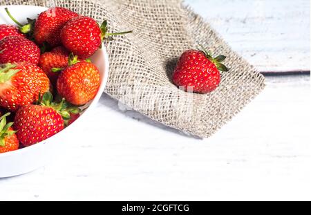 Berries of ripe strawberries lie in a white high plate, which stands on a light wooden background on a napkin made of burlap. Place for copy space Stock Photo
