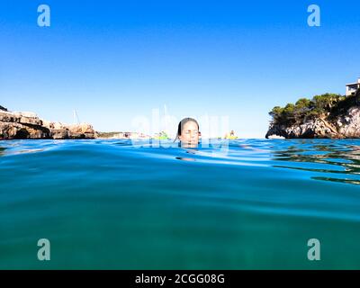 Young woman taking a bath in a beautiful cove in Majorca with only her head above the water and pleasure boats in the background. Holiday concept Stock Photo