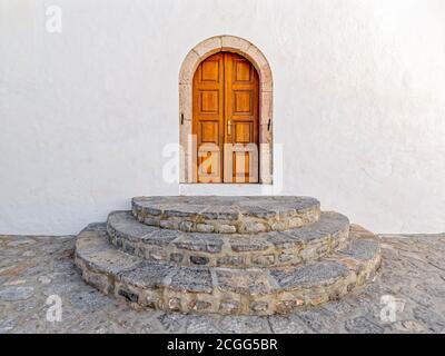 A natural wooden door with an arched stone frame on a white-washed wall and half-circle stone steps; Hydra island Greece. Stock Photo