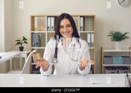 Smiling woman doctor looking at camera and showing something with hands in medical clinic Stock Photo