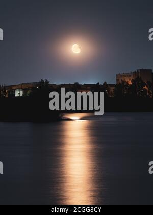 Bright moon shining on dark sky above lake water surface with big reflection and city silhouettes. Night time with large moon. Long exposure, vertical Stock Photo