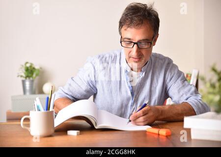 Smiling adult man with glasses studying on wooden table underlining in a book. Stock Photo