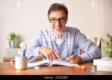 Smiling adult man with glasses studying on wooden table writing in a book. Stock Photo
