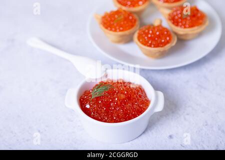 Red caviar (salmon caviar) in a white cup. In the background is a plate with tartlets and caviar. Selective focus, close up. Stock Photo