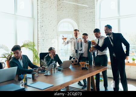 Watching football, drinking beer, cheering. Happy carefree colleagues having fun in office while their co-workers working hard and highly concentrated. Concept of fun, resting, professional occupation. Stock Photo