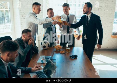 Watching football, drinking beer, cheering. Happy carefree colleagues having fun in office while their co-workers working hard and highly concentrated. Concept of fun, resting, professional occupation. Stock Photo
