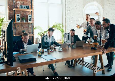 Watching football, drinking beer, cheering. Happy carefree colleagues having fun in office while their co-workers working hard and highly concentrated. Concept of fun, resting, professional occupation. Stock Photo
