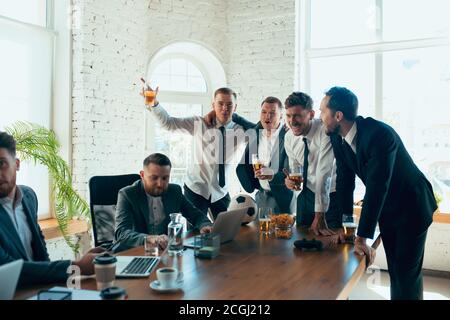 Watching football, drinking beer, cheering. Happy carefree colleagues having fun in office while their co-workers working hard and highly concentrated. Concept of fun, resting, professional occupation. Stock Photo