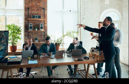 Watching basketball, drinking beer, cheering. Happy carefree colleagues having fun in office while their co-workers working hard and highly concentrated. Concept of fun, resting, professional occupation. Stock Photo