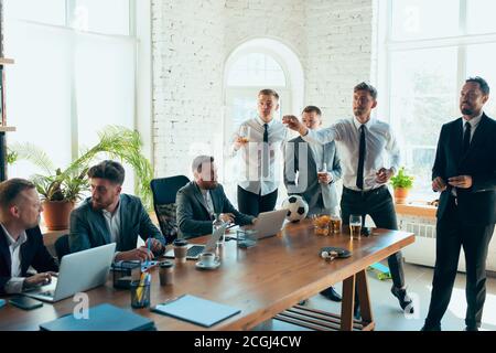 Watching football, drinking beer, cheering. Happy carefree colleagues having fun in office while their co-workers working hard and highly concentrated. Concept of fun, resting, professional occupation. Stock Photo