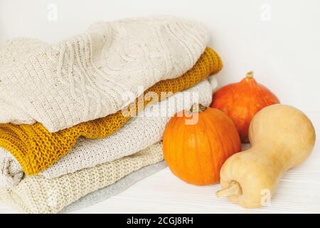 Cozy knitted sweaters and pumpkins on white background. Hello fall! Stylish white and yellow sweaters stack and orange pumpkins with space for text. H Stock Photo