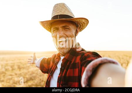 Image of excited adult man in straw hat taking selfie photo while standing at cereal field Stock Photo