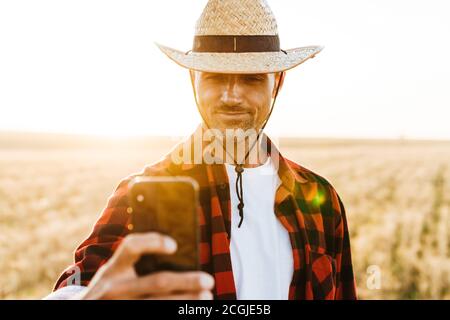 Image of pleased adult man in straw hat taking selfie on mobile phone at cereal field Stock Photo
