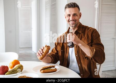 Image of grey-haired joyful man drinking soda and eating sandwiches while have lunch in white kitchen Stock Photo