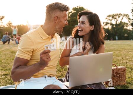 Portrait of joyful middle-aged couple man and woman holding credit card while using laptop computer during picnic in summer park Stock Photo