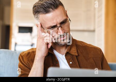 Image of perplexed adult man in eyeglasses working with laptop while sitting on couch at home Stock Photo