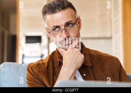 Image of thinking adult man in eyeglasses working with laptop while sitting on couch at home Stock Photo