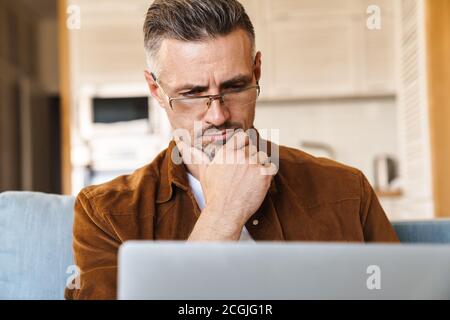 Image of perplexed adult man in eyeglasses working with laptop while sitting on couch at home Stock Photo