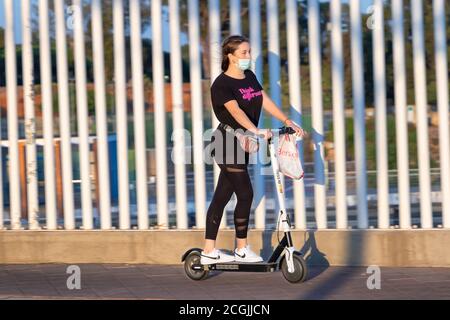 Huelva, Spain - September 10, 2020:  Young woman riding an electric scooter by the sidewalk wearing a protective mask due to coronavirus covid-19 Stock Photo
