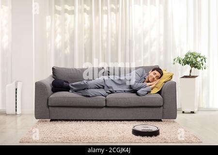Robotic vacuum cleaner vacuuming the carpet while a man in pajamas is sleeping on a sofa in a living room Stock Photo