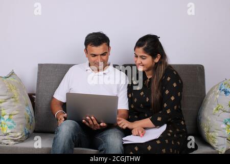 People, money and finances concept. Young Indian woman sitting near her husband on sofa, working with documents and laptop. Stock Photo