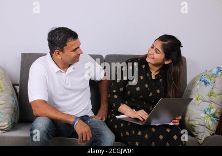 People, money and finances concept. Young Indian woman sitting near her husband on sofa, working with documents and laptop. Stock Photo