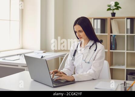 Young female doctor typing on laptop keyword surfing net in modern hospital office Stock Photo