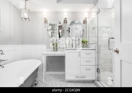 A beautiful white bathroom with a stand alone clawfoot bathtub with chrome hardware. Flowers are on the white marble vanity counter top. Stock Photo