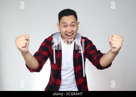 Successful young Asian man shows winning gesture, celebrating victory. Hands raised above with expressive omg wow happy face Stock Photo