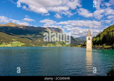 The ancient submerged bell tower of Lake Resia in Val Venosta, South Tyrol, Italy, emerges from the water against a beautiful blue sky with white clou