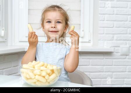 4 years old girl sits at the kitchen table with corn sticks in a glass cup, in the hands of a child corn sticks and smiles Stock Photo