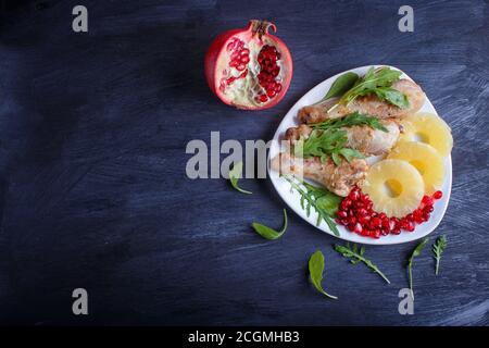 Fried chicken legs with rucola, pineapple and pomegranate seeds on black wooden background, copy space, top view. Stock Photo
