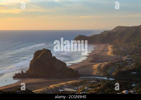 Piha, New Zealand, at sunset. Lion Rock towers above the black sand beach. In the background is Te Waha Point Stock Photo
