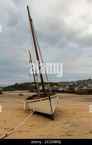 A white small wooden fishing boat is stranded on the beach at low tide. This old sail boat is tilted on one side. City scapeof the Cornish Fishing tow Stock Photo