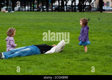 Paris/France 06/13/2010: A father is lying on grass in an outdoor park. He is trying to rest or sleep but his two little daughters do not leave him al Stock Photo