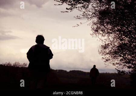 Silhouette of two men walking away in a rural area at sunset on a cloudy day. The man behind is in focus and has winter clothings. His dishevelled hai Stock Photo