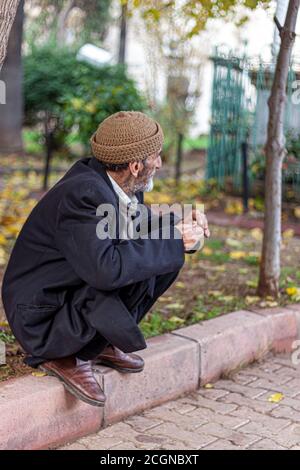 Adana, Turkey, 12/31/2009: An elderly man wearing traditional hat, winter coat, and leather shoes is squatting on the edge of a sidewalk. He is lookin Stock Photo