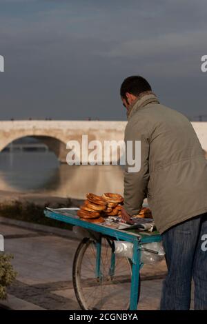 A young man is selling simit (Turkish sesame bagel) on a wooden food cart in the street. Historic Stone bridge over Seyhan river in Adana is in the ba Stock Photo