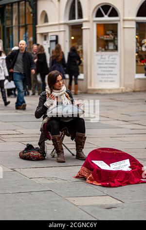 Bath, UK, 03/06/2010: A young woman musician is sitting on a foldable stool in a crowded street and playing hang instrument. This street performer is Stock Photo
