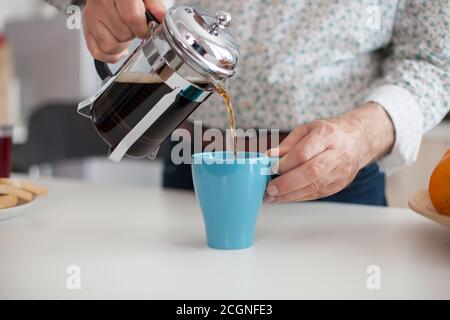 Senior man pouring hot coffee from french press during breakfat in kitchen. Elderly person in the morning enjoying fresh brown cafe espresso cup caffeine from vintage mug, filter relax refreshment Stock Photo