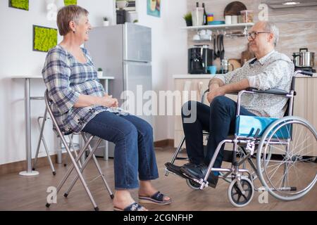 Old woman and her disabled husband in wheelchair chatting in kitchen.Elderly person having a conversation with husband in kitchen. Living with disabled person with walking disabilities Stock Photo