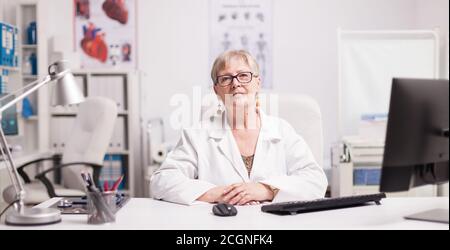 Elderly aged woman doctor in hospital cabinet wearing white coat. Stock Photo