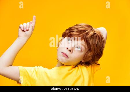 Expressive red-haired boy front view in a yellow t-shirt points up with index finger close-up, Copy space Stock Photo