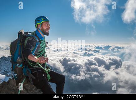 Smiling bearded climber in a safety harness, helmet, and on body wrapped climbing rope sitting at 3600m altitude on a cliff and looking at  picturesqu Stock Photo