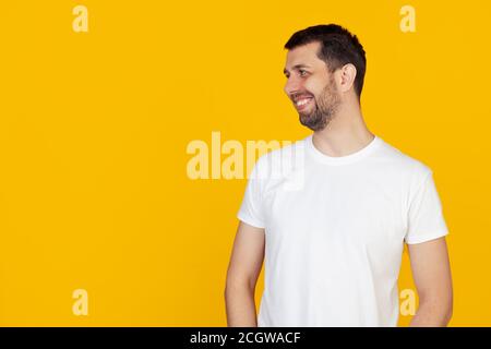 Young man with a beard in a white t-shirt looking to the side with a smile on his face, natural expression. Laughs confidently, stands on isolated yellow background. Stock Photo