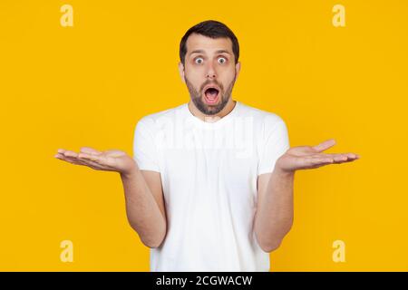Young man with a beard in a white T-shirt spreading his arms out, shocked by the unexpected expression, hesitates to say, the answer has no idea. I don't know stands on isolated yellow background. Stock Photo