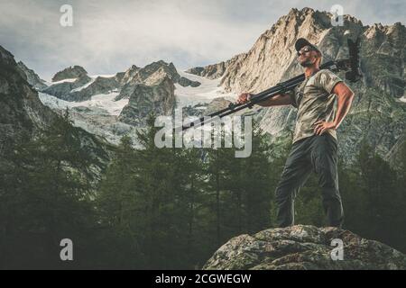 Caucasian Commercial Nature Photographer in His 40s with Professional Photo Equipment on His Shoulder. Digital Camera on a Tripod. Alpine Landscape in Stock Photo