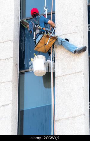 An unidentified window washer working high up on a building. He sits on a very small platform suspended by rope and washes a windows with a large brus Stock Photo